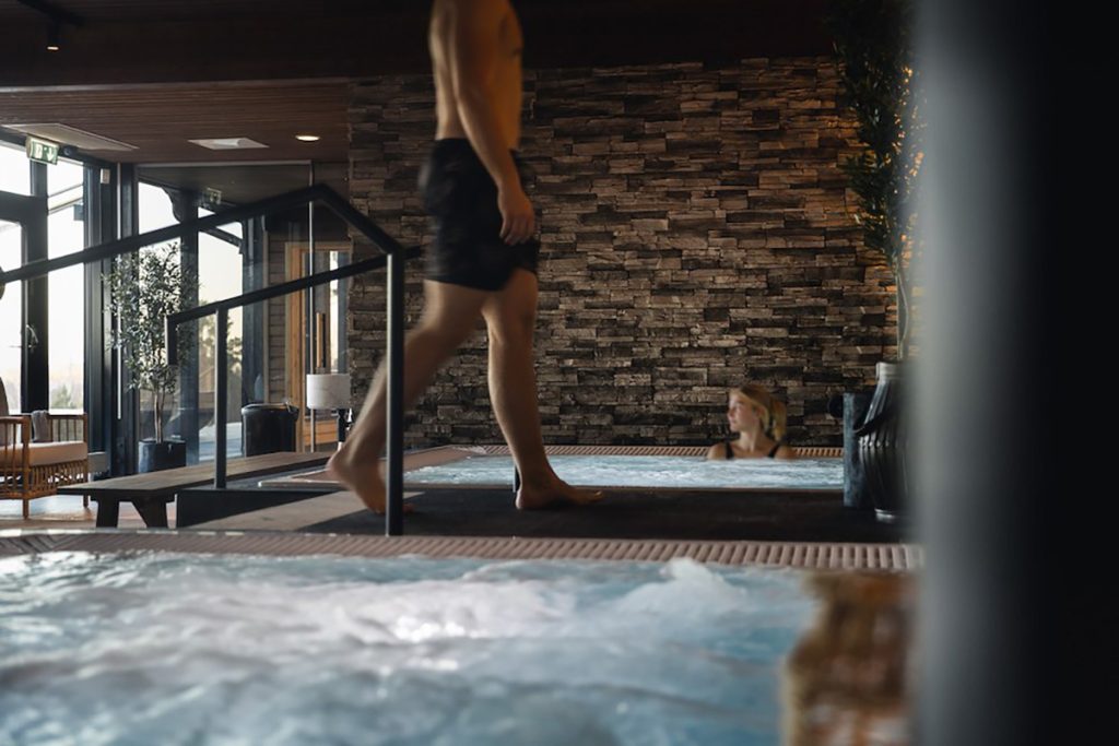 En man i badbyxor går bredvid en inomhuspool med bubblande vatten, medan en kvinna kopplar av i vattnet i närheten. Spaavdelningen har en vägg av sten och en mjuk inomhusbelysning.