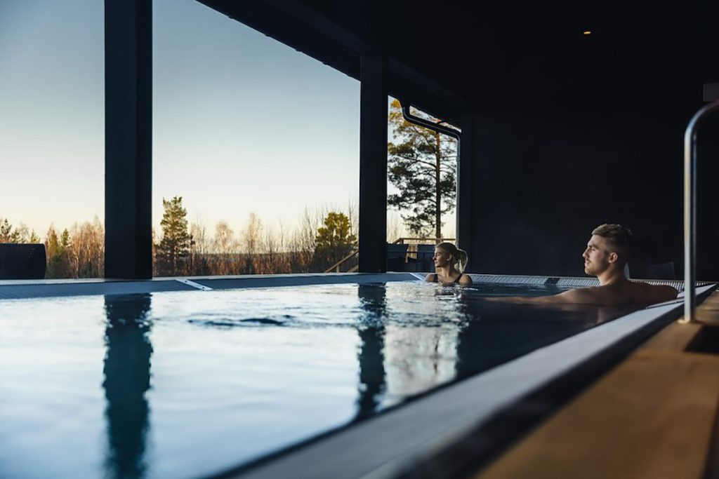 Två personer kopplar av i en inomhuspool med stora fönster och tittar ut på träd och en klar himmel under solnedgången eller soluppgången. Det varma ljuset kontrasterar mot den mörka interiören.